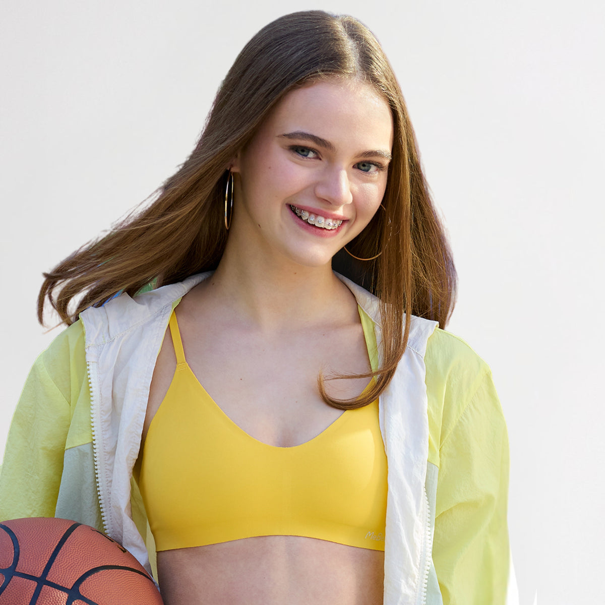 Teen Girl in yellow bra and green jacket holding a basketball against a light background