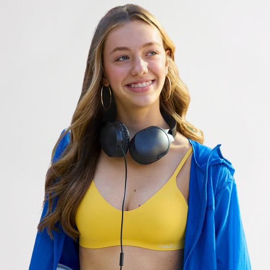 Teen Girl wearing a yellow bra and blue jacket with headphones around her neck on a light gray background