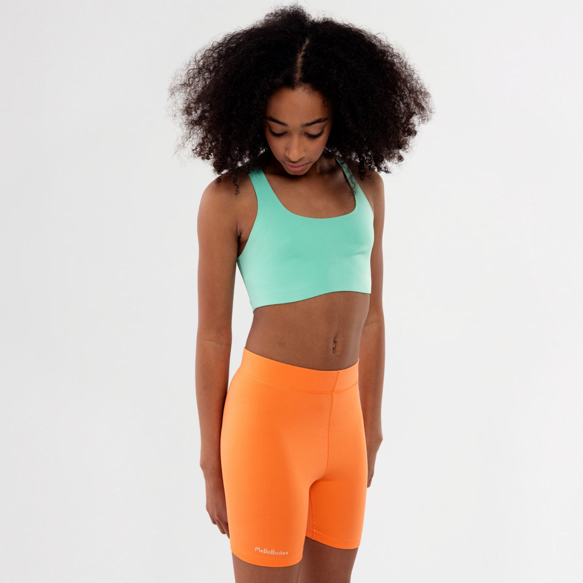 Teen Girl wearing a green sports bra and orange shorts on a white background