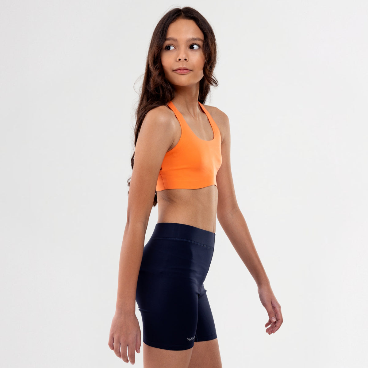 Teen Girl wearing an orange sports bra and black shorts on a white background
