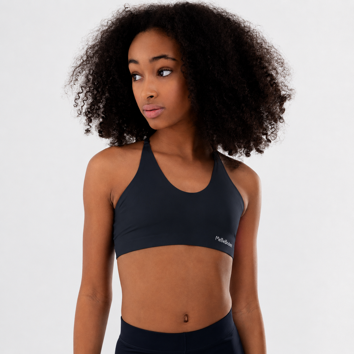 A tween girl with curly dark hair wearing a navy athletic sports bra with a small white logo on the side, photographed against a clean light grey studio background.