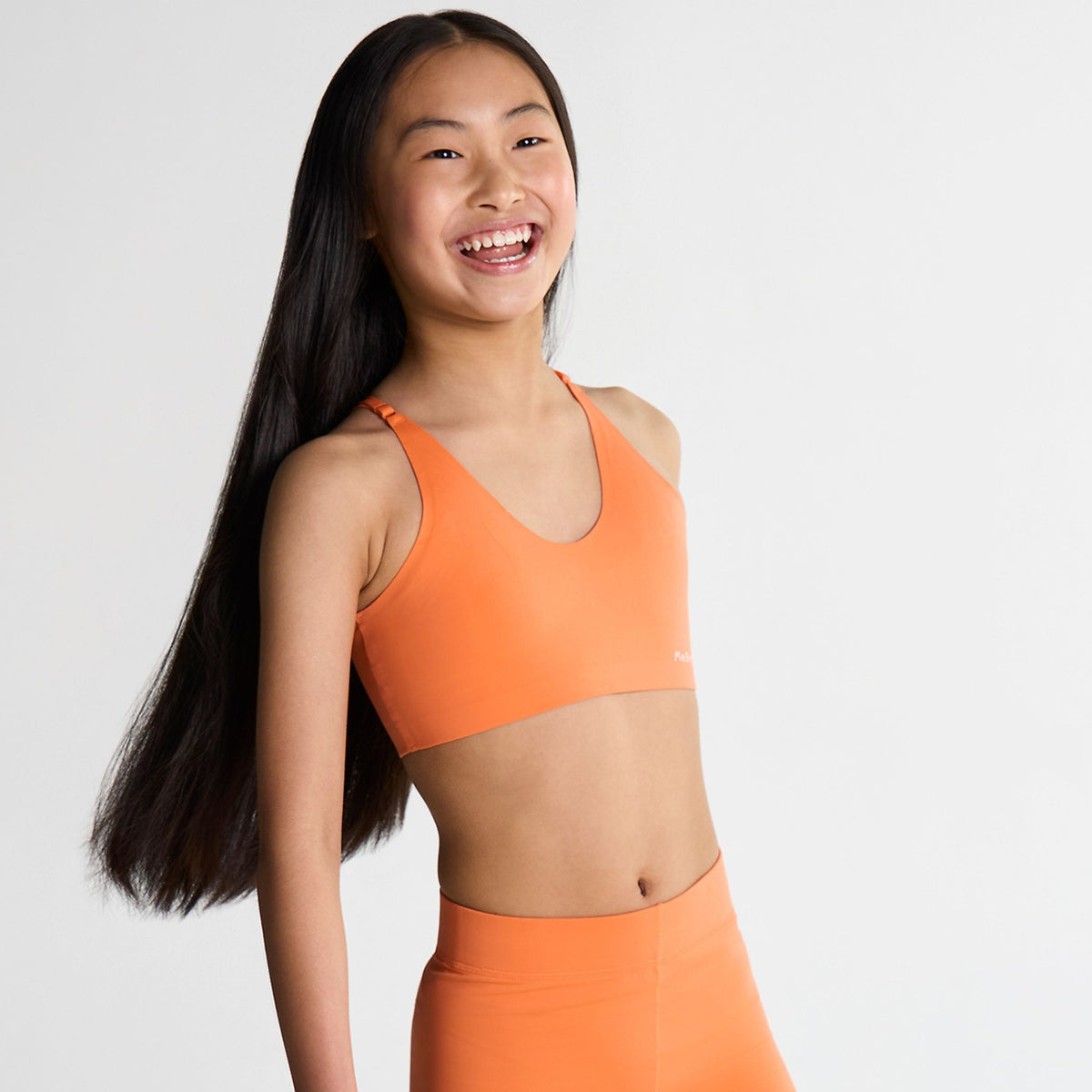Tween Girl wearing an orange sports bra and leggings on a light gray background