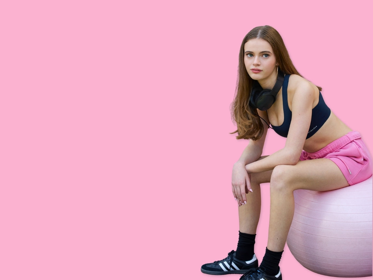 A teen girl in athletic wear sits on a pink exercise ball against a solid pink background. She is wearing a mebebodee branded navy sports bra, pink shorts, black socks, and black-and-white sneakers.