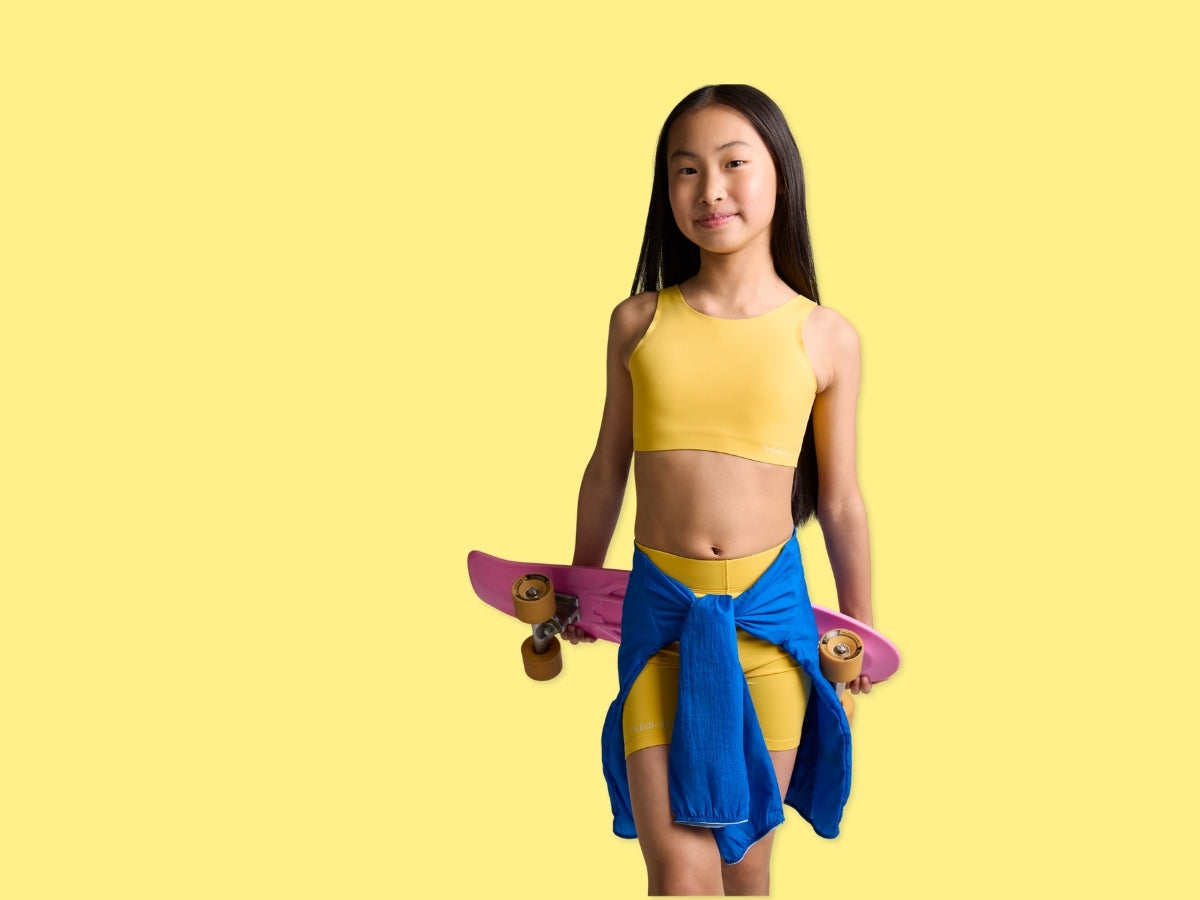 A tween girl with long dark hair stands against a solid yellow background, holding a pink skateboard. She is wearing a yellow mebebodee branded crop top and matching yellow shorts, with a blue shirt tied around her waist.